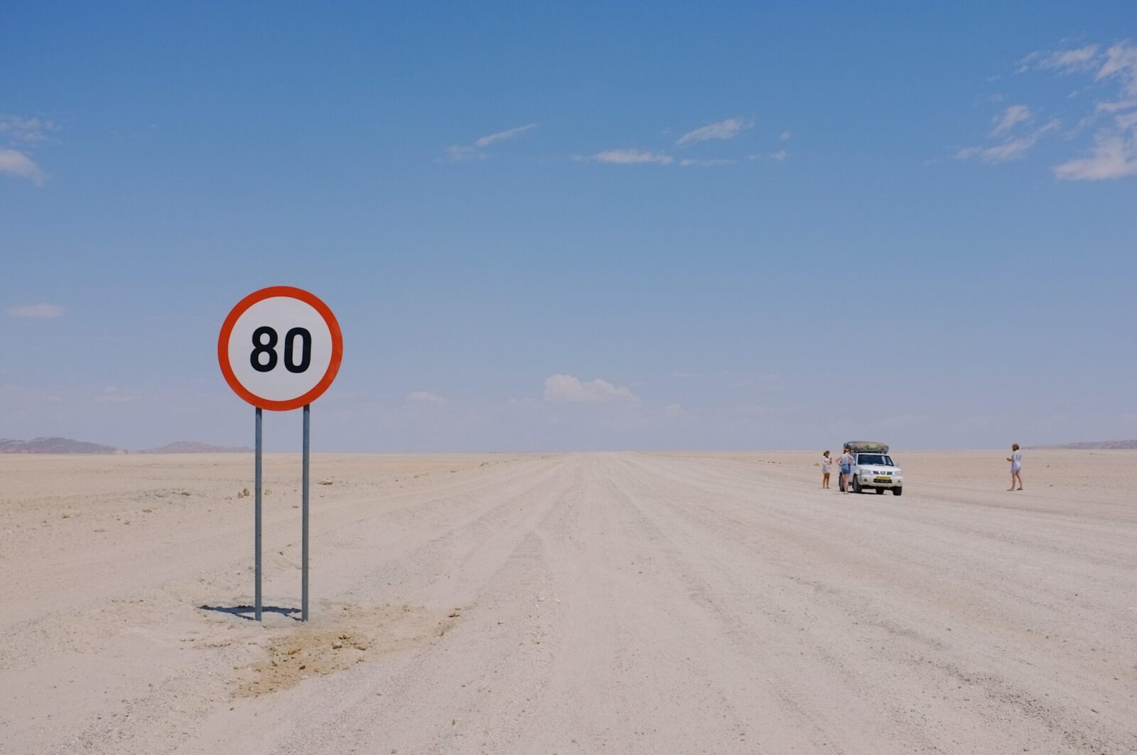 A distant desert road sign displaying 80km speed limit with a car and travelers visible.